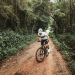 Man in cycling gear biking down a dirt road through a lush forest.